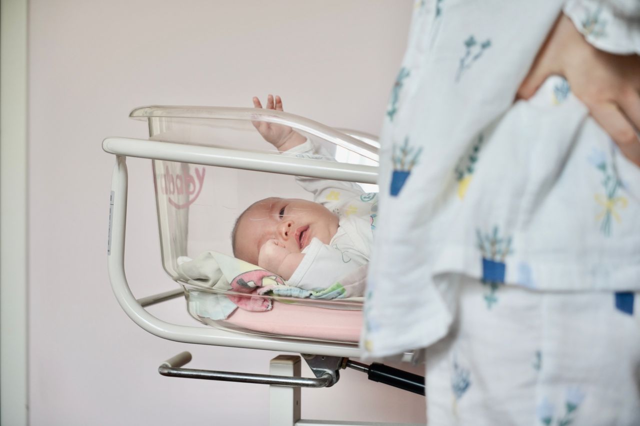 A tender scene of a newborn baby lying in a hospital bassinet with the mother standing close by.