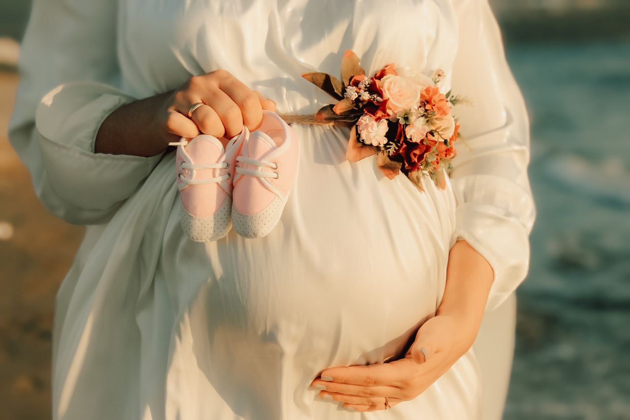 Expecting mother in white dress holding pink baby shoes at the beach, symbolizing a new life.