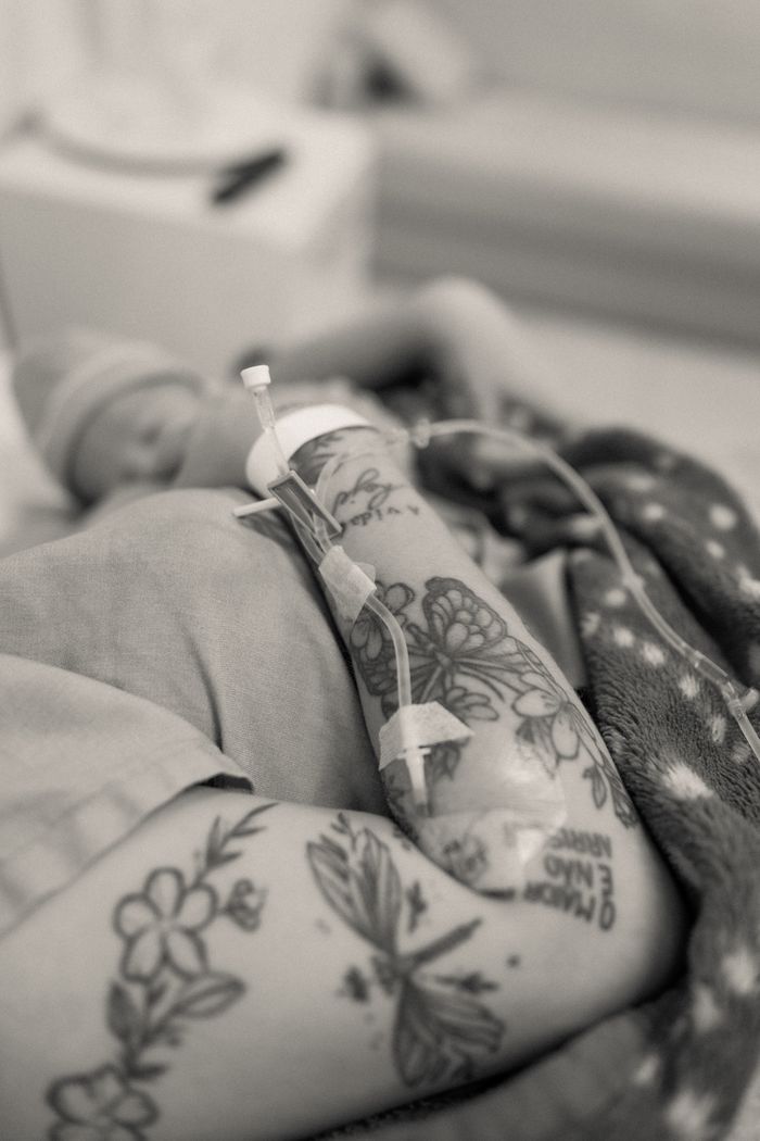 A close-up of a tattooed parent's arm with IV next to a newborn baby in a hospital setting.