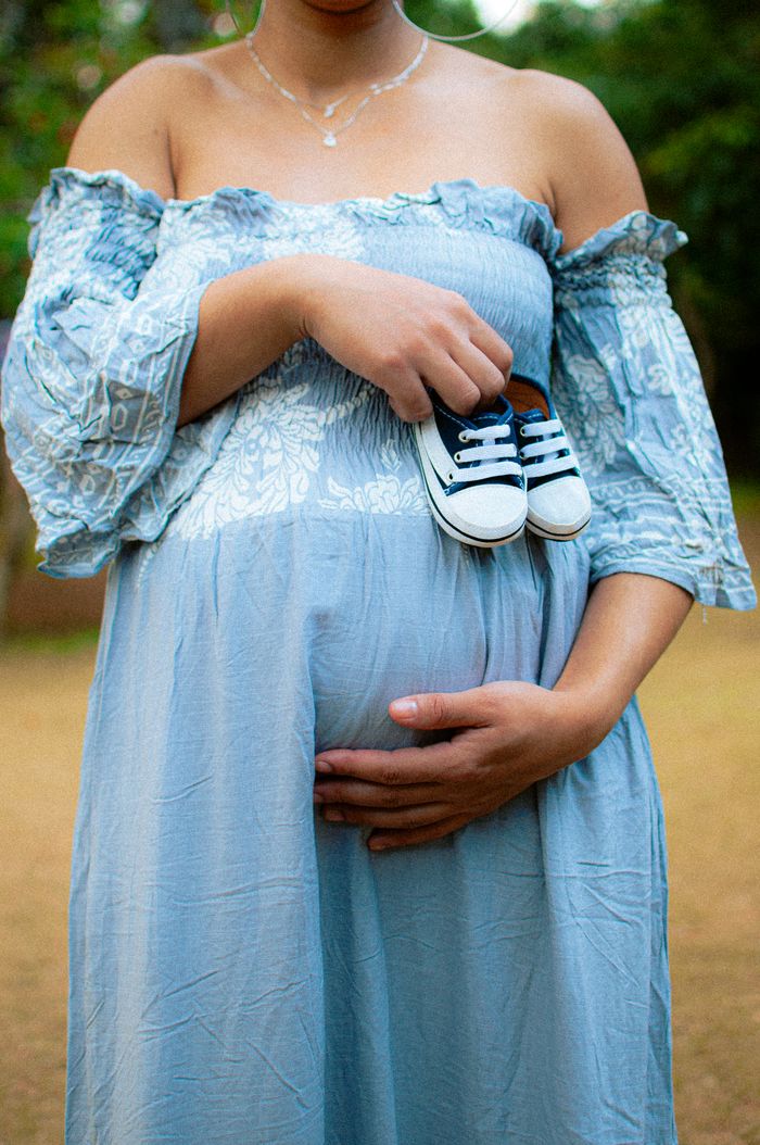 Pregnant woman in a blue dress holds baby shoes outdoors, symbolizing joy and anticipation.