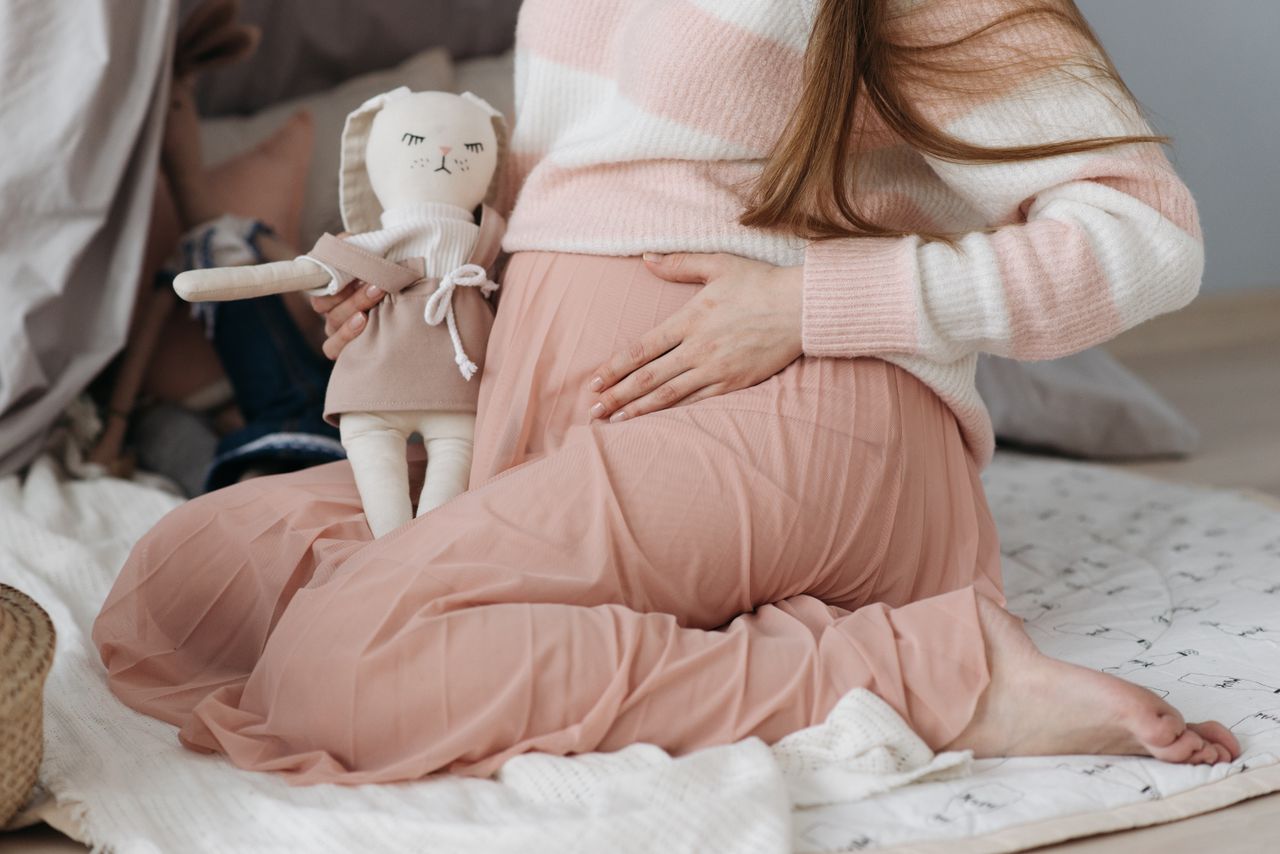 Expectant mother sitting on the floor holding a soft toy, wearing pink and white cozy attire.